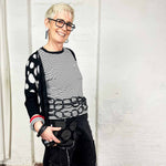 Woman with blond cropped hair wearing black square glasses and wearing a rubber chain necklace as a belt around her waist.