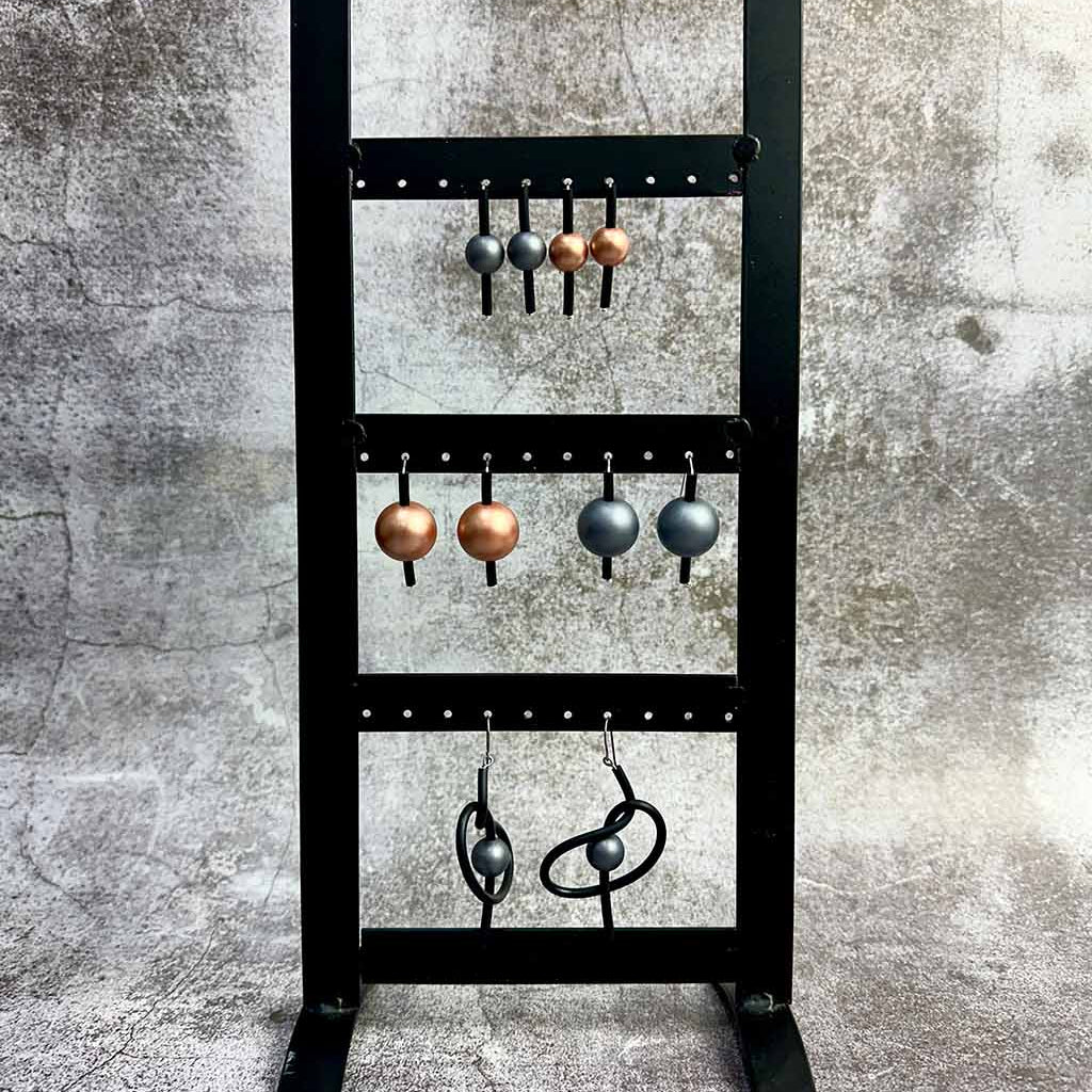 A black metal stand displaying six pairs of metallic earrings on hooks against a textured grey background.