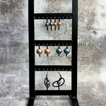 A black metal stand displaying six pairs of metallic earrings on hooks against a textured grey background.