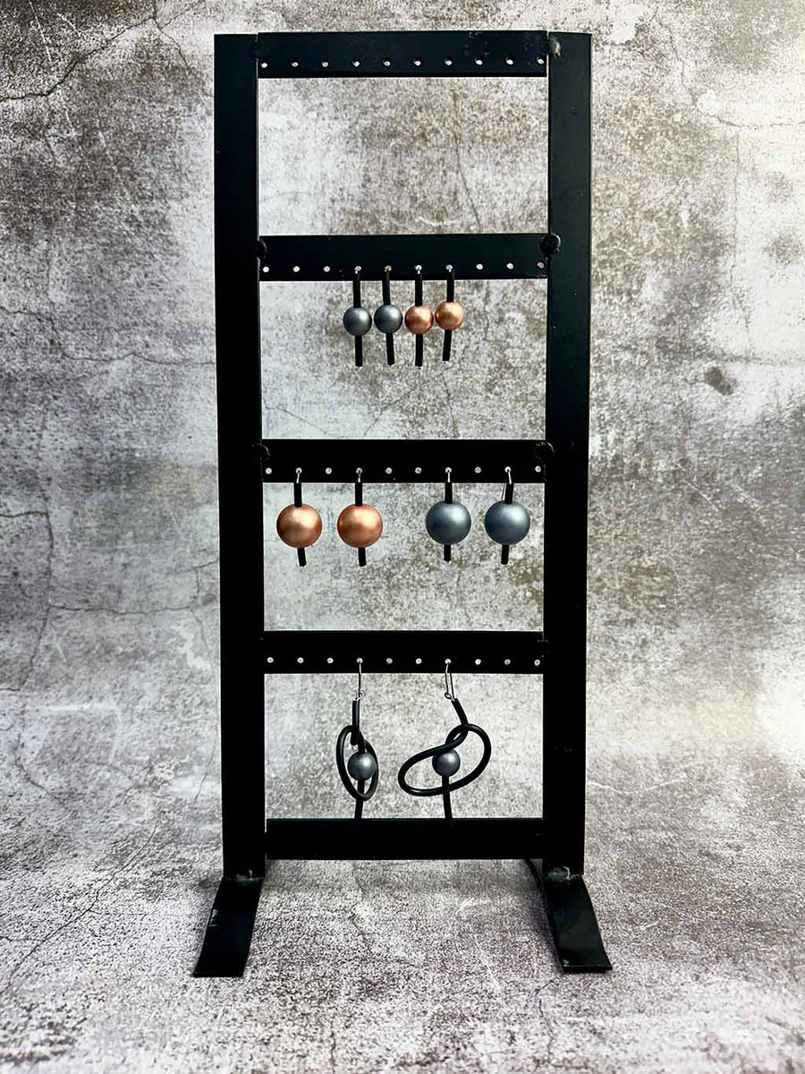 A black metal stand displaying six pairs of metallic earrings on hooks against a textured grey background.