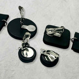 A set of black and silver metal clip-on accessories arranged on a textured white background, including round and square black discs with shiny silver metal clips.