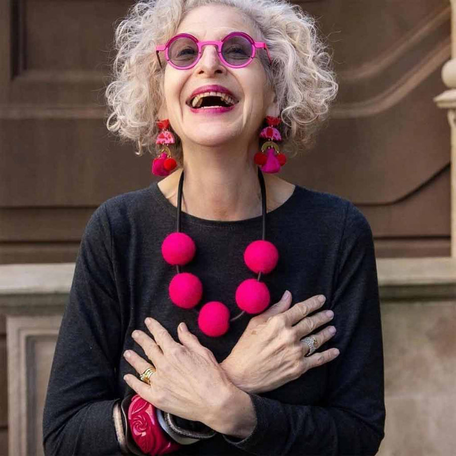 Woman with round pink glasses wearing big dangle earrings and necklace with large pink felt balls